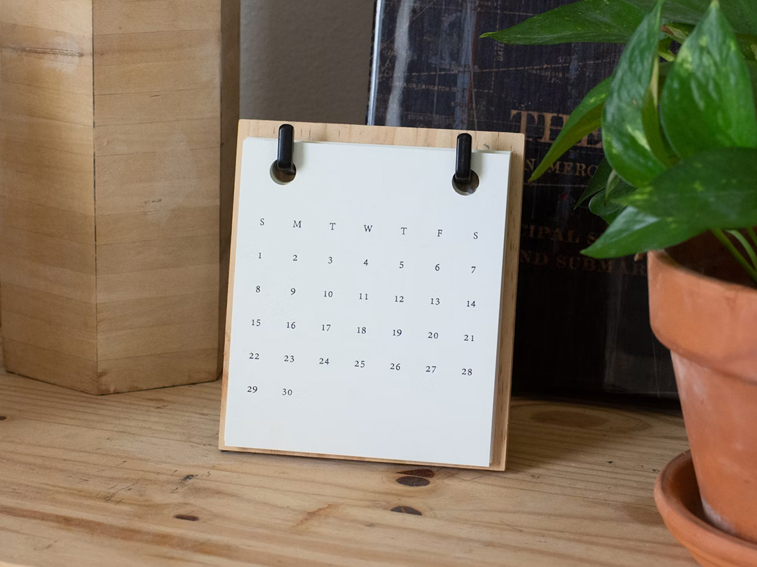 Paper calendar stand on a wooden table, next to a potted plant.